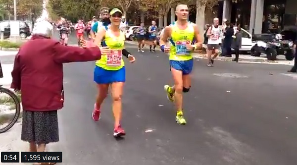 screenshot of video shared by Age UK of older lady standing next to runners in a race. She holds out her arm to get high fives from friendly runners.